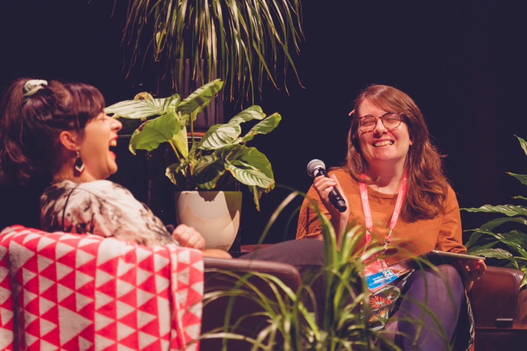 Lisa Blakie (left) and Jess Woodward (right) seated in armchairs, surrounded by plants and laughing together during a Q&A session at Play by Play 2021.